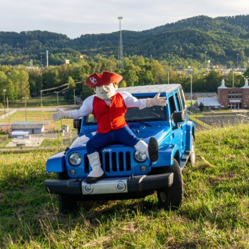 Mascot sitting on a car with a campus building in the backround