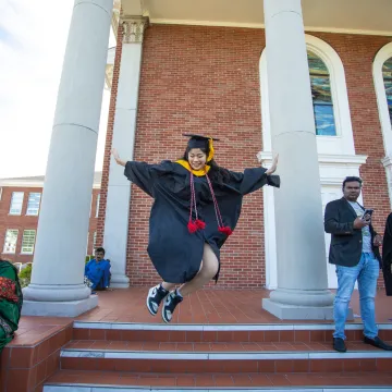 A graduate leaping for joy in front of a building. 