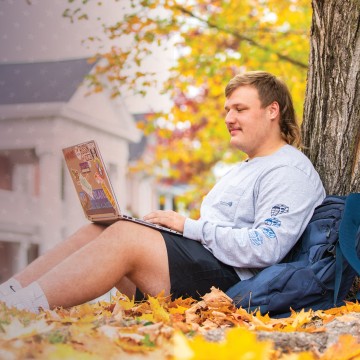 A student studies under a tree amongst fallen leaves on campus.