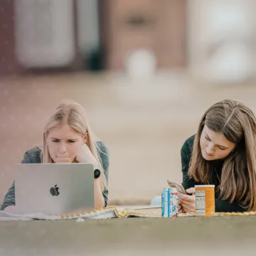 Two females lay on a blanket on campus and study. 