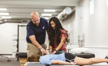 A physical therapy student is participating in a clinical exercise checking the back of another student with an instructor watching. 