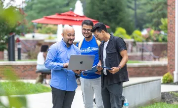 Three male students look at their computer while standing in the campus quad. 