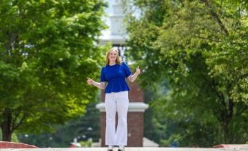 A female student in a blue shirt gives the thumbs up in front of the campus clock tower. 