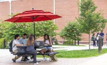 Four students sit at a table with their computers studying outside. 