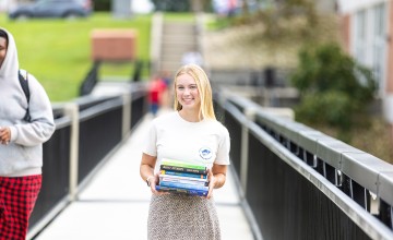 A female student stands on the campus viaduct holding a stack of textbooks. 