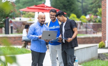 Three male students, one in a Cumberlands shirt, look at a laptop together in the campus quad. 