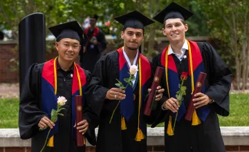 Three male students hold a rose and their diploma following graduation. 
