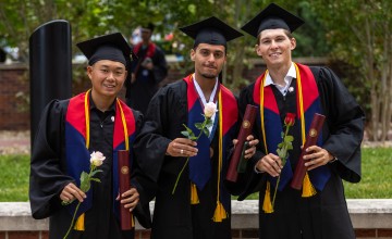 Three male students hold a rose and their diploma following graduation. 