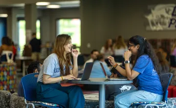 Two female students sit with their laptops at a round table in the student center. 