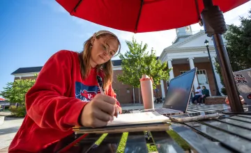 A female student sits at a table outside on campus and writes in her notebook. 