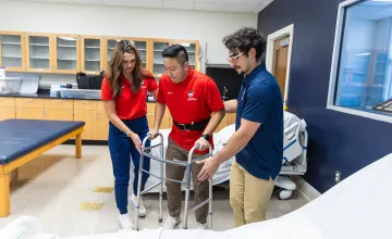 Two medical science students assist another student with a walking assistant during a classroom exercise. 