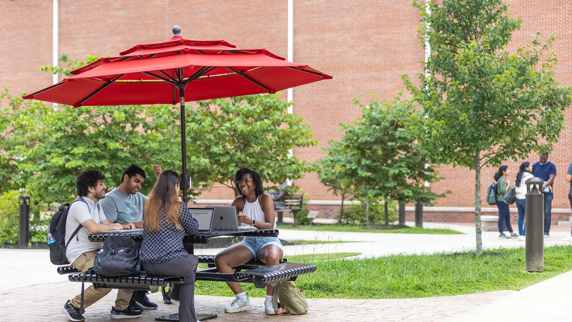 Four students sit at a table with their computers studying outside. 