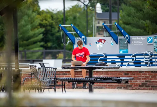 Male student sitting on a brick wall outside, holding a laptop.
