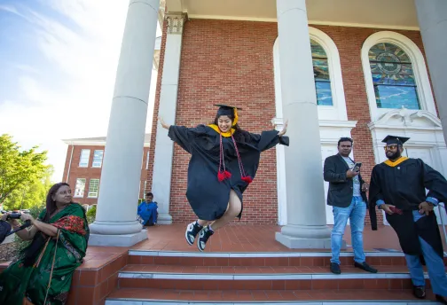 A graduate leaping for joy in front of a building. 