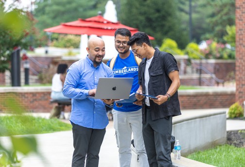 Three male students look at their computer while standing in the campus quad. 