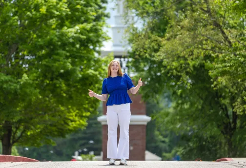 A female student in a blue shirt gives the thumbs up in front of the campus clock tower. 