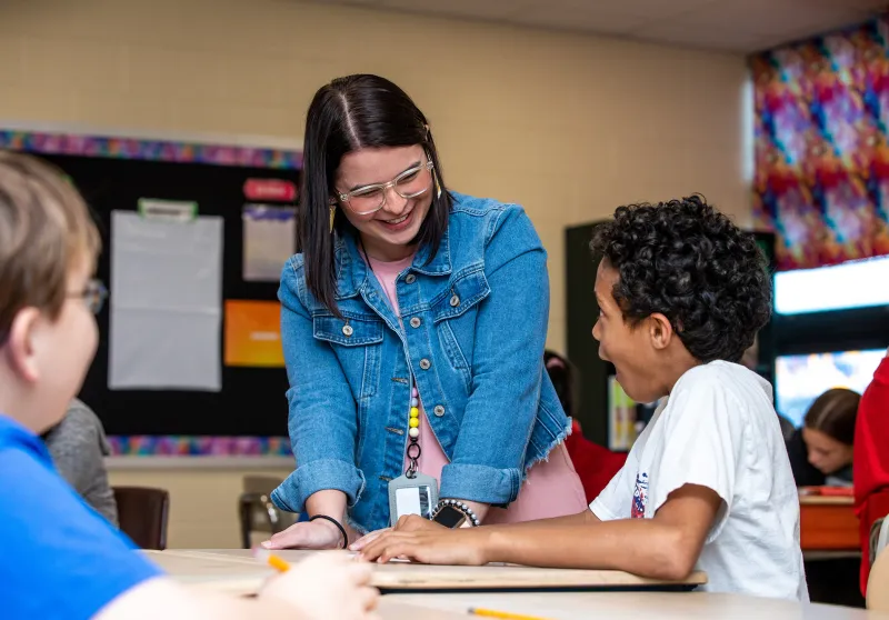  A teacher talking to students sitting at a desk while in a classroom. 