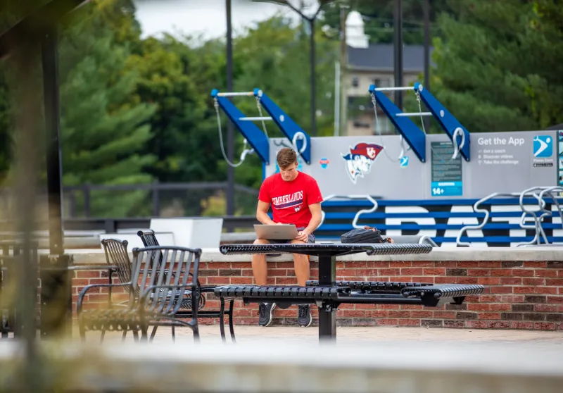 Male student sitting on a brick wall outside, holding a laptop.