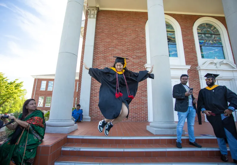 A graduate leaping for joy in front of a building. 