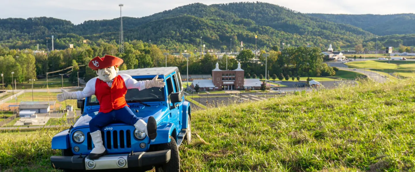 Mascot sitting on a car with a campus building in the backround
