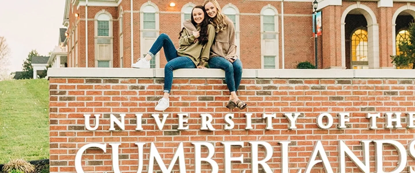 Two students posing for a picture seated on top of the University of the Cumberlands campus sign