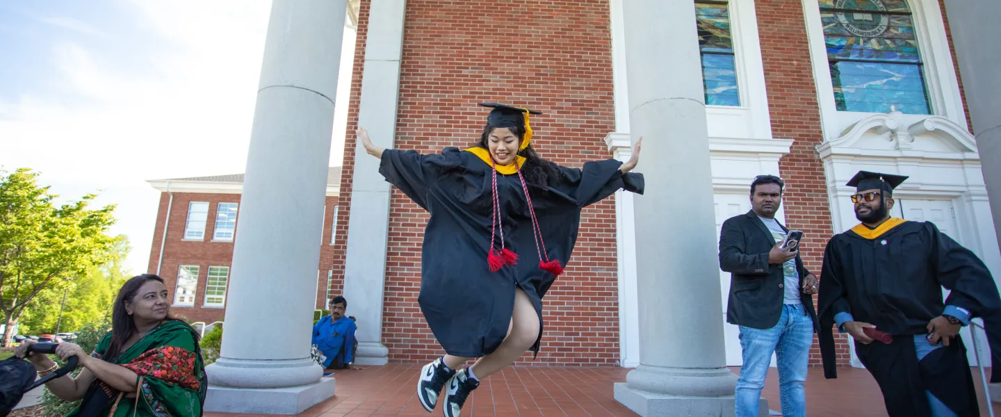 A graduate leaping for joy in front of a building. 
