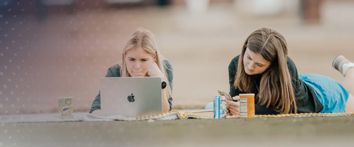 Two females lay on a blanket on campus and study. 