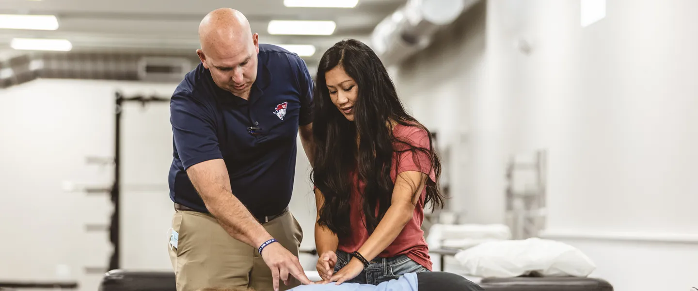 A physical therapy student is participating in a clinical exercise checking the back of another student with an instructor watching. 