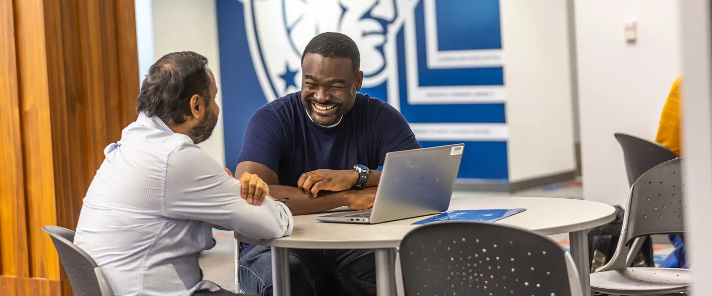 Two male students chat at a table with their computer inside the Patriot Success Center. 