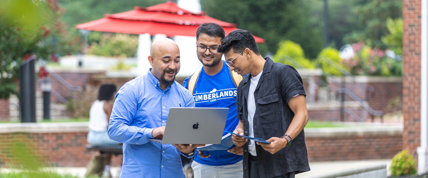 Three male students look at their computer while standing in the campus quad. 