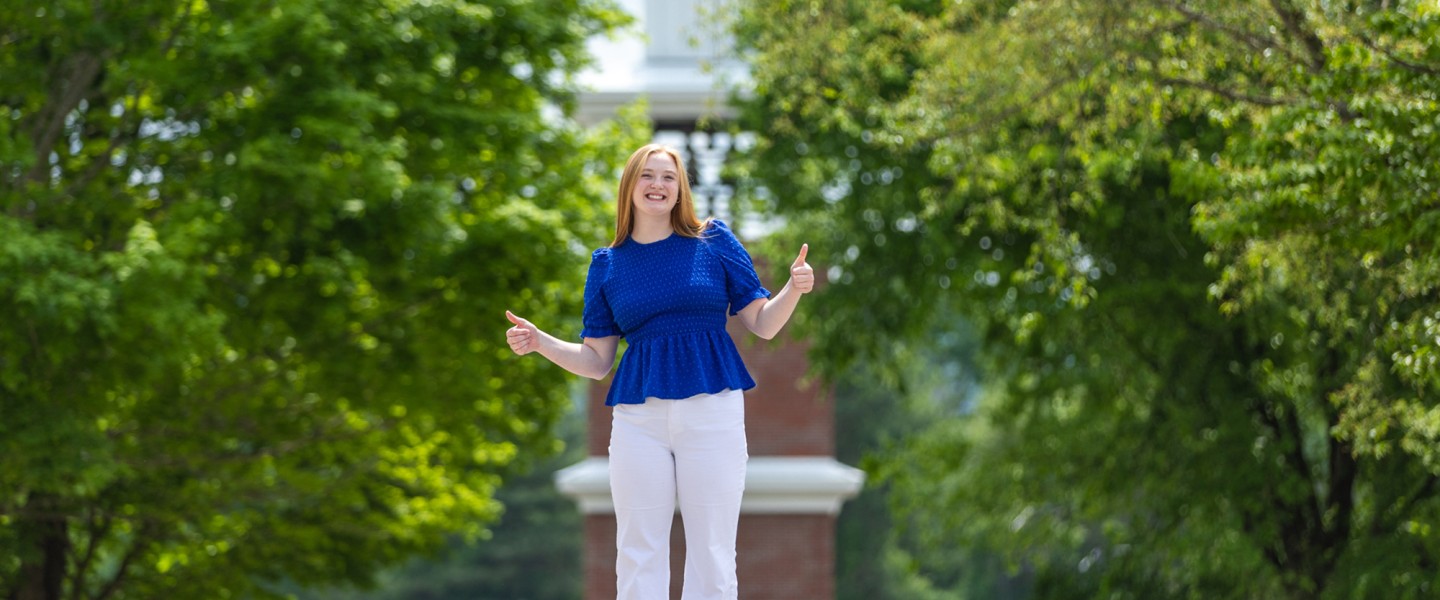 A female student in a blue shirt gives the thumbs up in front of the campus clock tower. 