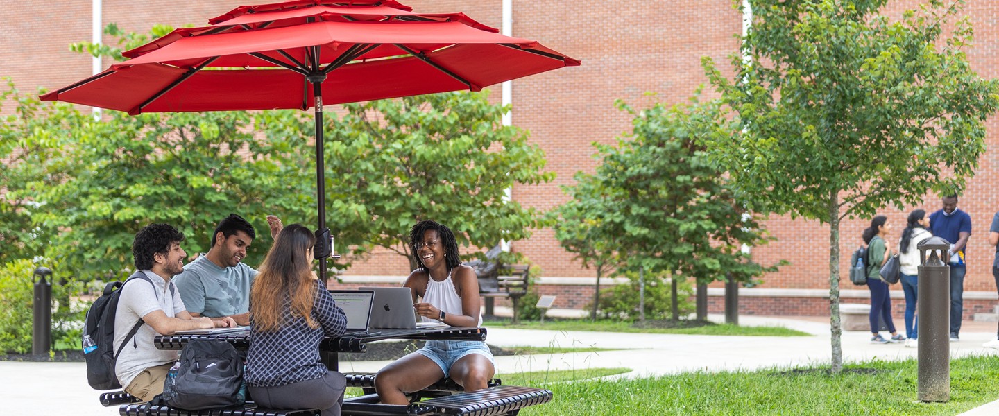 Four students sit at a table with their computers studying outside. 