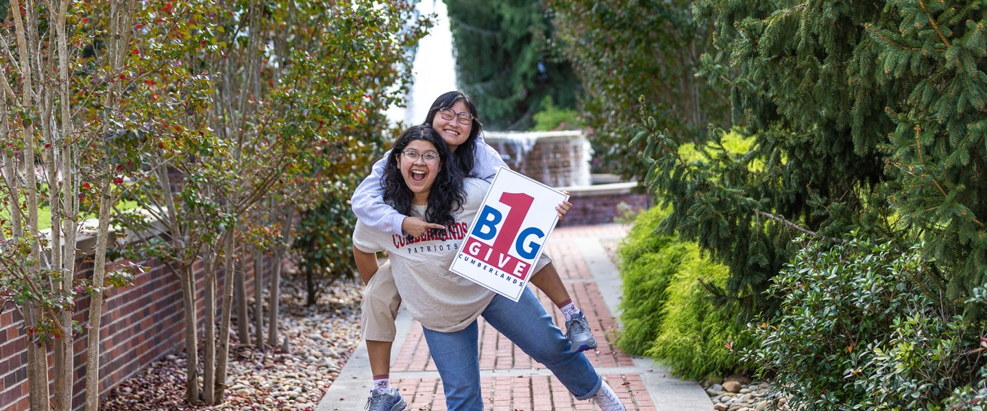 Two female students promote the One Big Give event while standing in front of the fountain. 