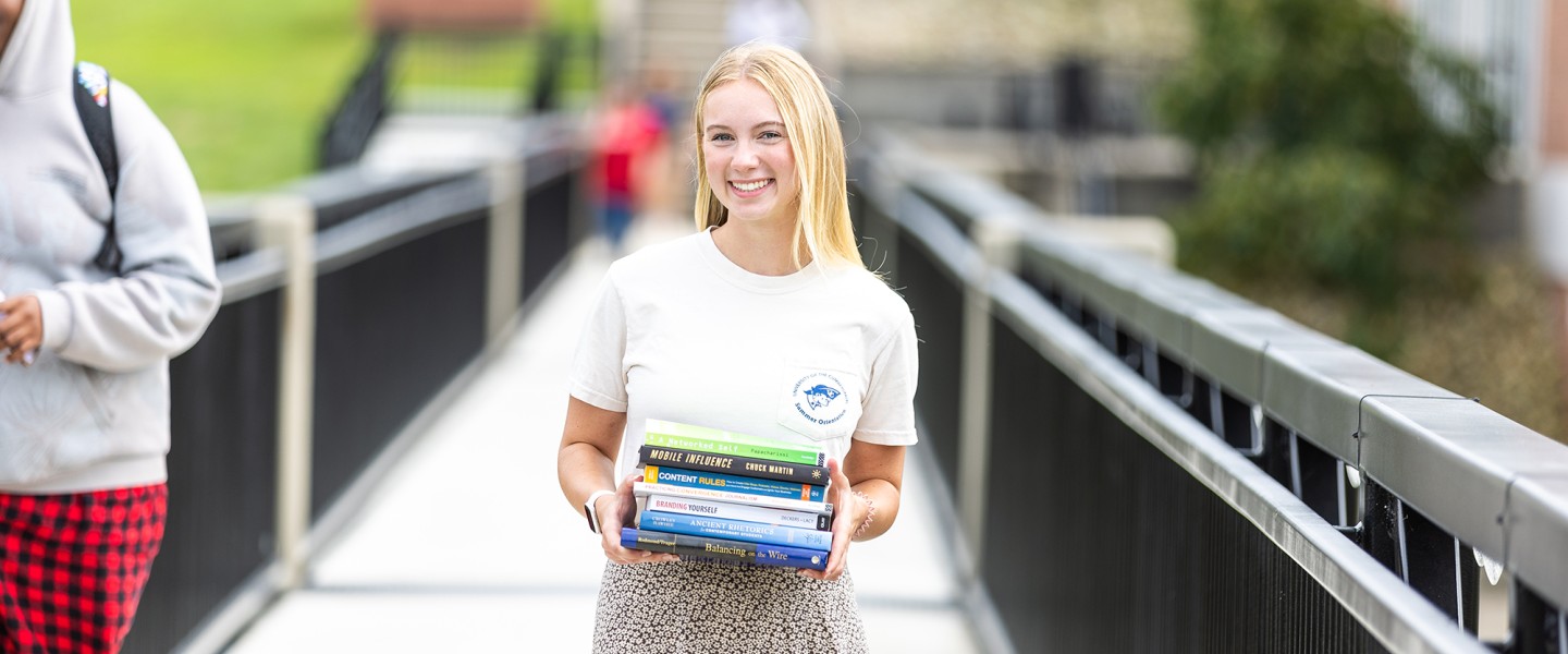 A female student stands on the campus viaduct holding a stack of textbooks. 