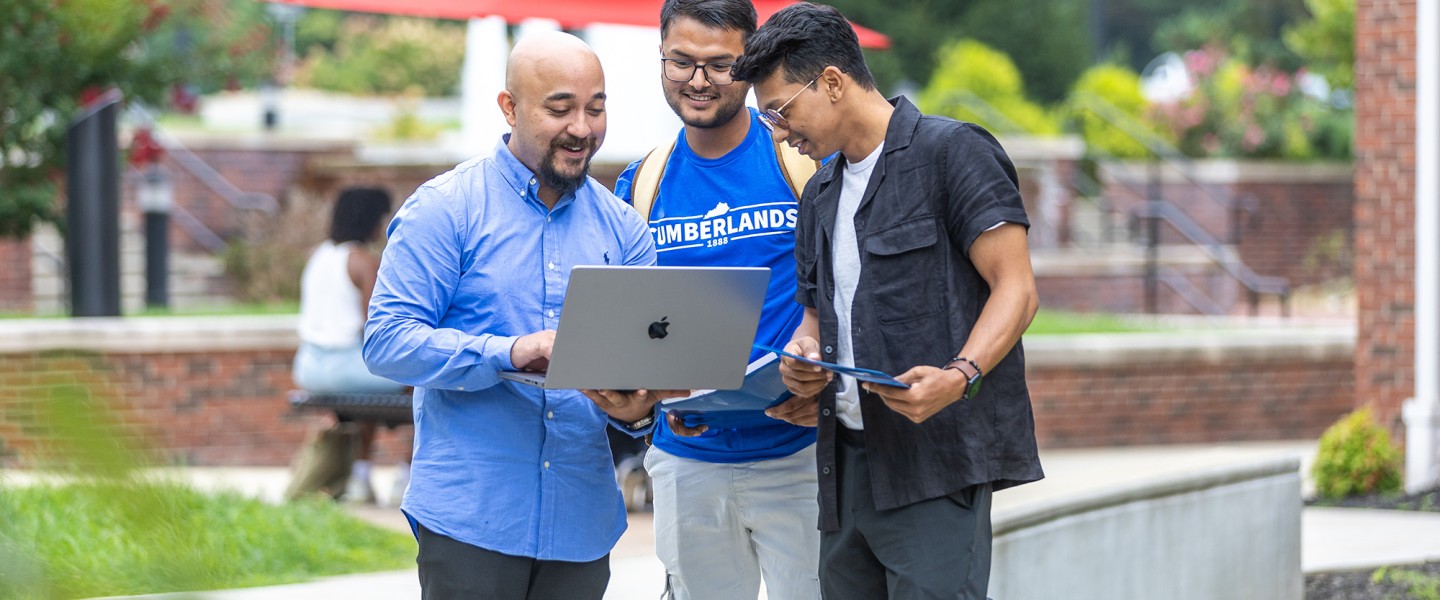 Three male students, one in a Cumberlands shirt, look at a laptop together in the campus quad. 