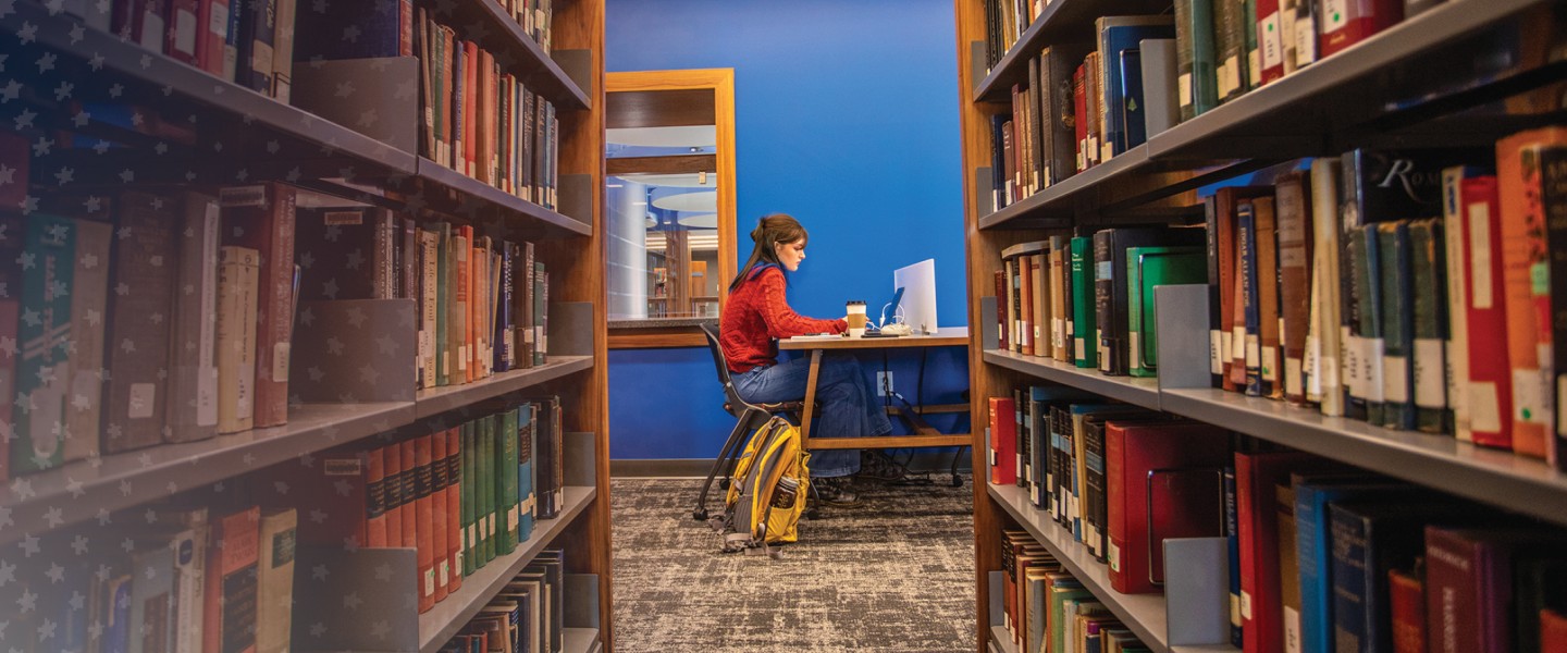 A female student sits at a booth in the library studying with shelves of books in the foreground.