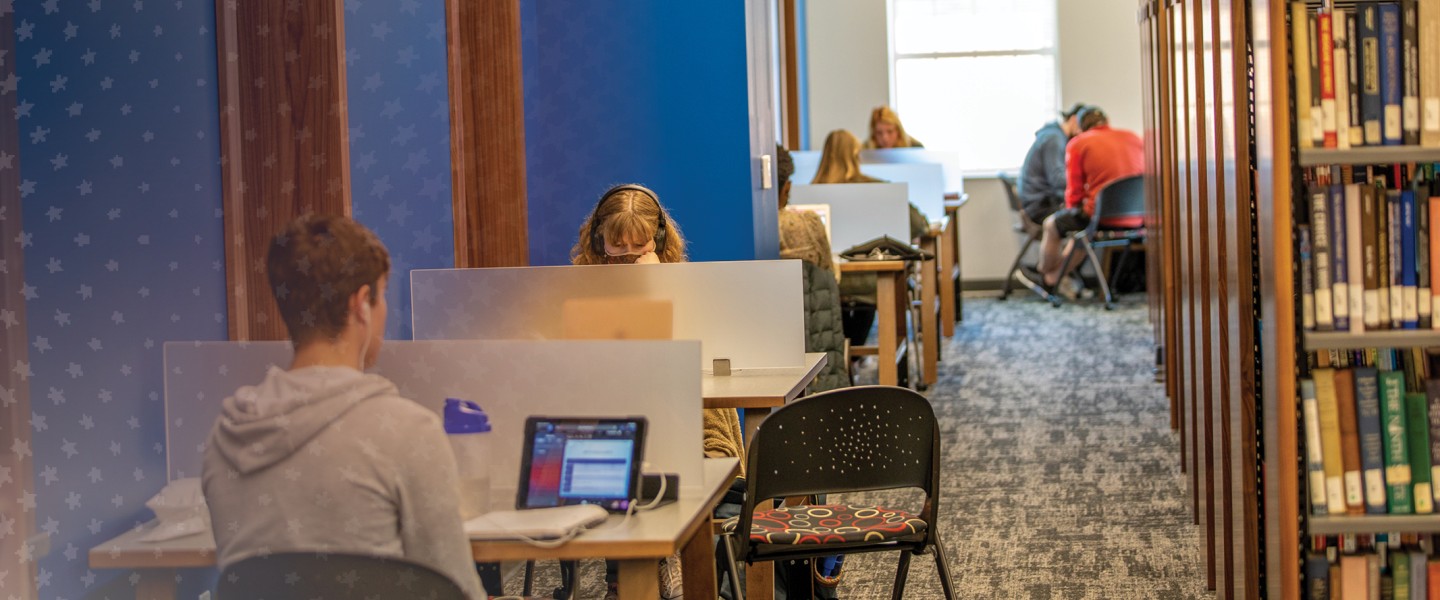 Several students sit at study desks inside the campus library with book shelves to their right. 