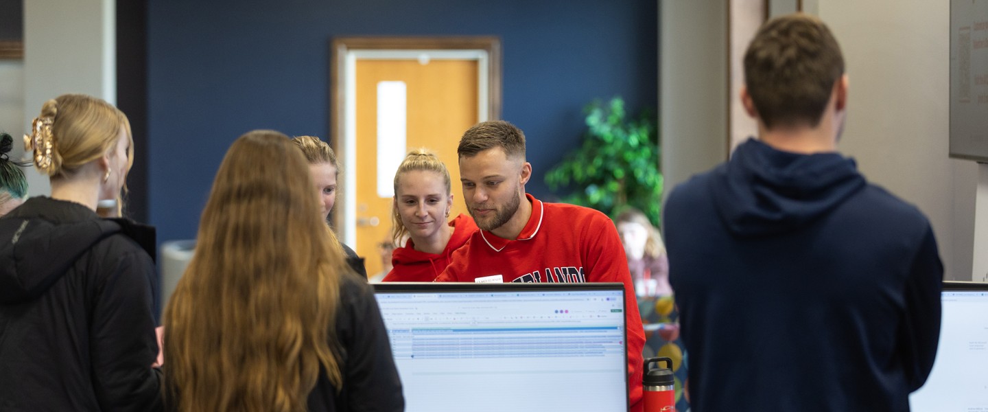 A group of students watch a male student demonstrate using AI during an info session in the student center. 