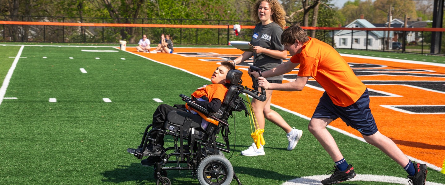 A male high school student, being coached by a Cumberlands student volunteer, pushes a child in a wheel chair during the 2026 Special Olympics.