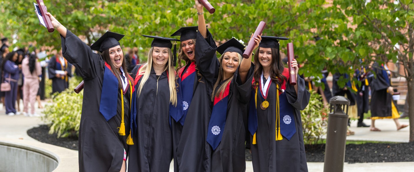 A group of five females in regalia celebrate with their diplomas following commencement services. 