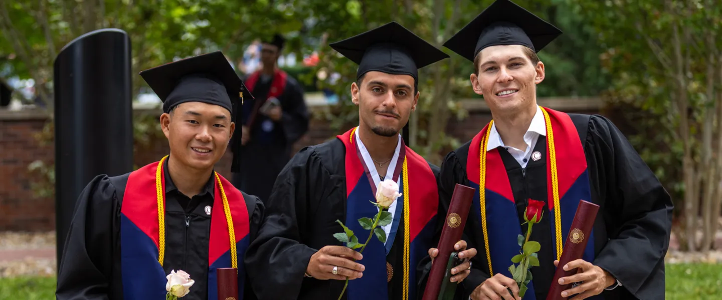 Three male students hold a rose and their diploma following graduation. 
