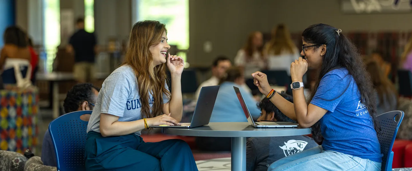 Two female students sit with their laptops at a round table in the student center. 