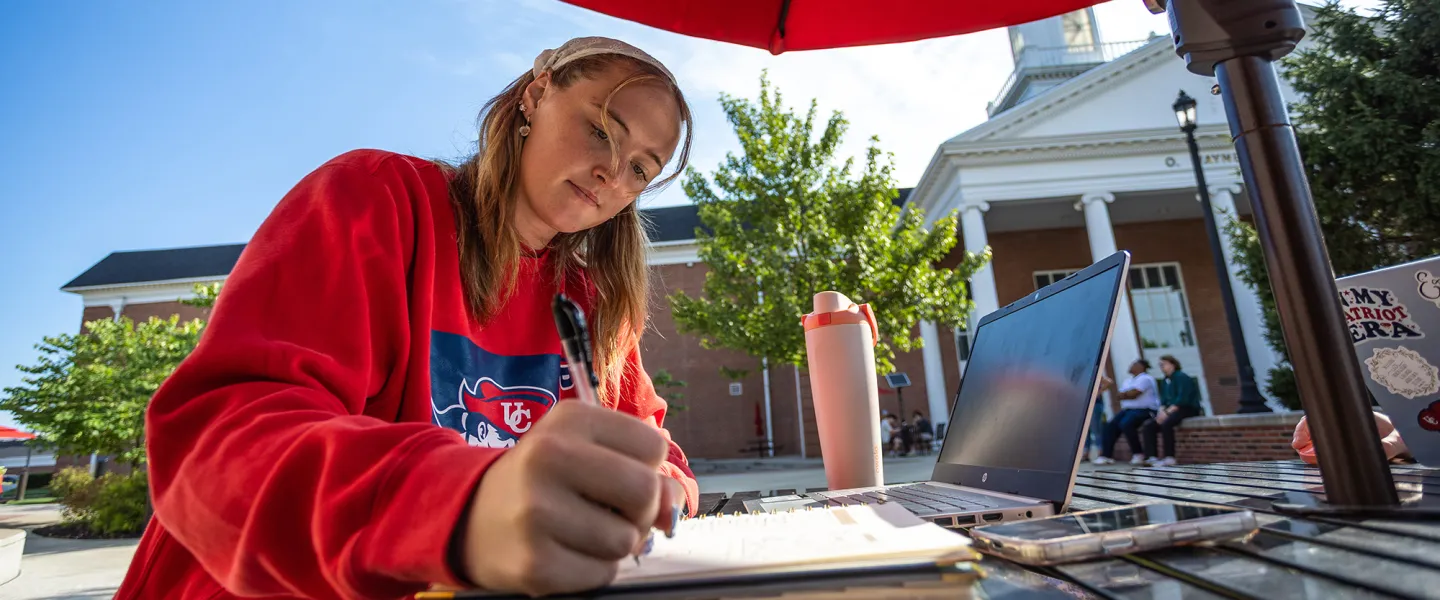 A female student sits at a table outside on campus and writes in her notebook. 