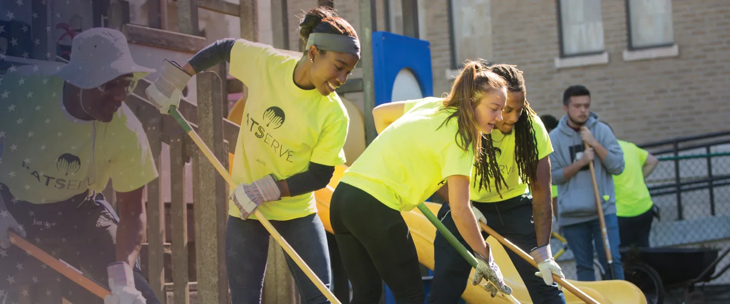 Two male and two female students use rakes to clean weeds from a flowerbed during a service event. 