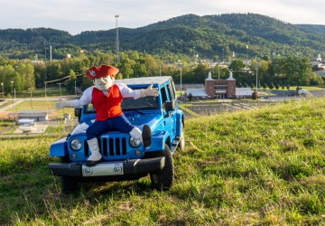 Mascot sitting on a car with a campus building in the backround