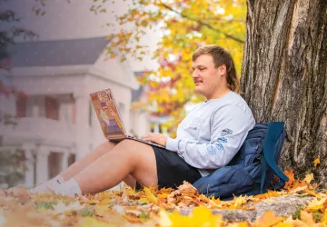A student studies under a tree amongst fallen leaves on campus.