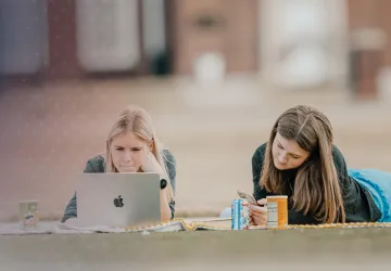 Two females lay on a blanket on campus and study. 
