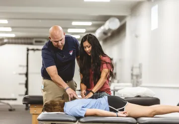 A physical therapy student is participating in a clinical exercise checking the back of another student with an instructor watching. 