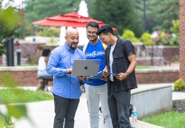Three male students look at their computer while standing in the campus quad. 