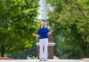 A female student in a blue shirt gives the thumbs up in front of the campus clock tower. 